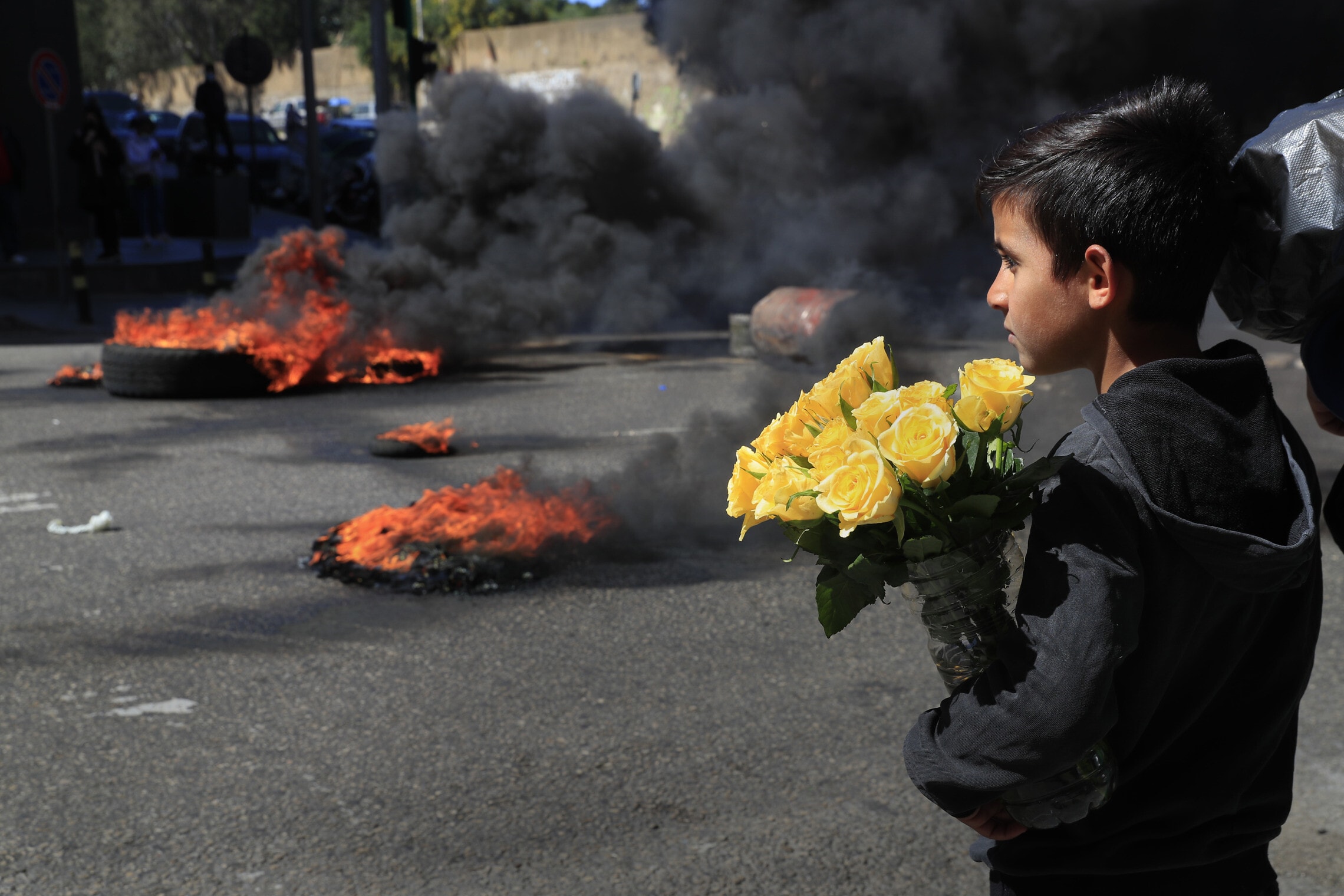 ילד מוכר פרחים מביט לעבר המפגינים ששורפים צמיגים במחאה על עליית המחירים והתרסקות הכלכלה הלבנונית, ביירות, 16 במרץ 2921 (צילום: AP Photo/Hussein Malla)