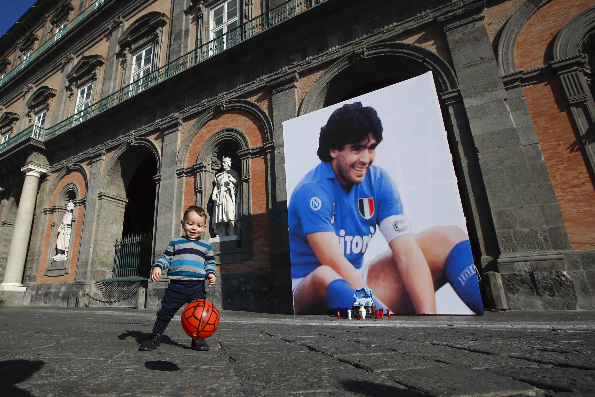 פעוט משחק עם כדור ליד תמונה של מראדונה בנאפולי, 2020 (צילום: AP Photo/Alessandra Tarantino)