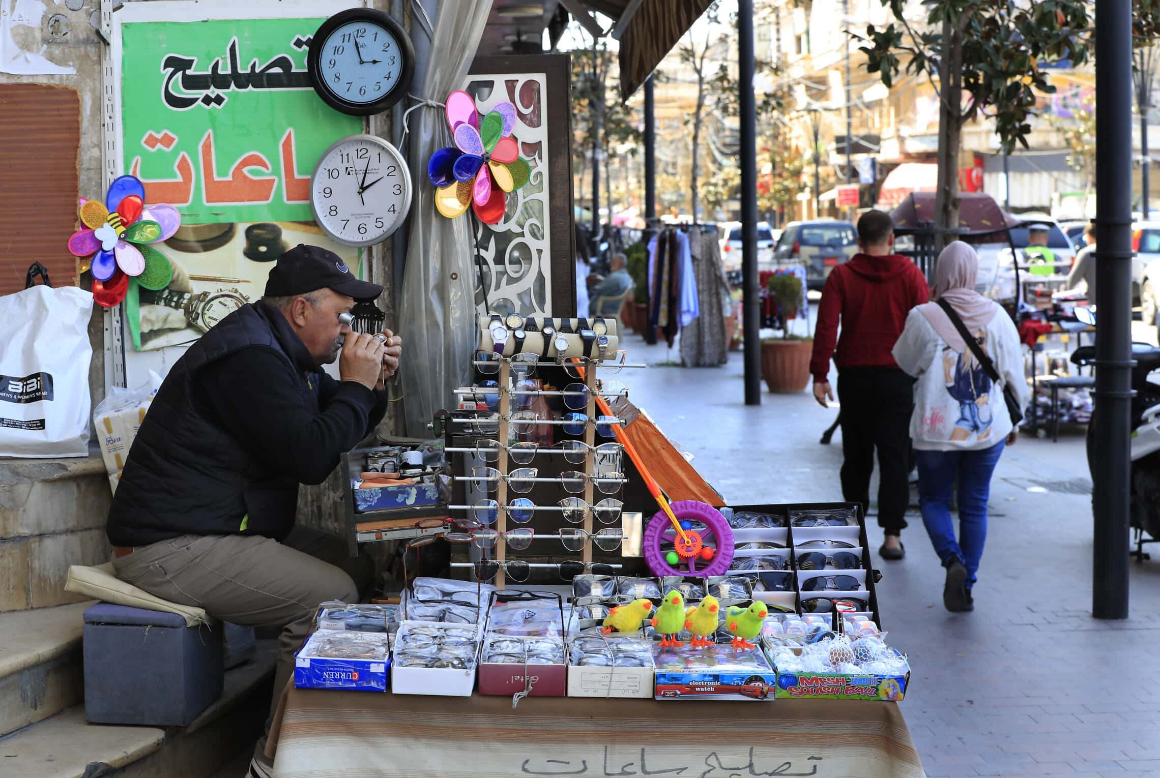 רוכל יושב ליד שני שעונים המראים את הזמנים השונים בלבנון, 27 במרץ 2023 (צילום: AP Photo/Mohammed Zaatari)
