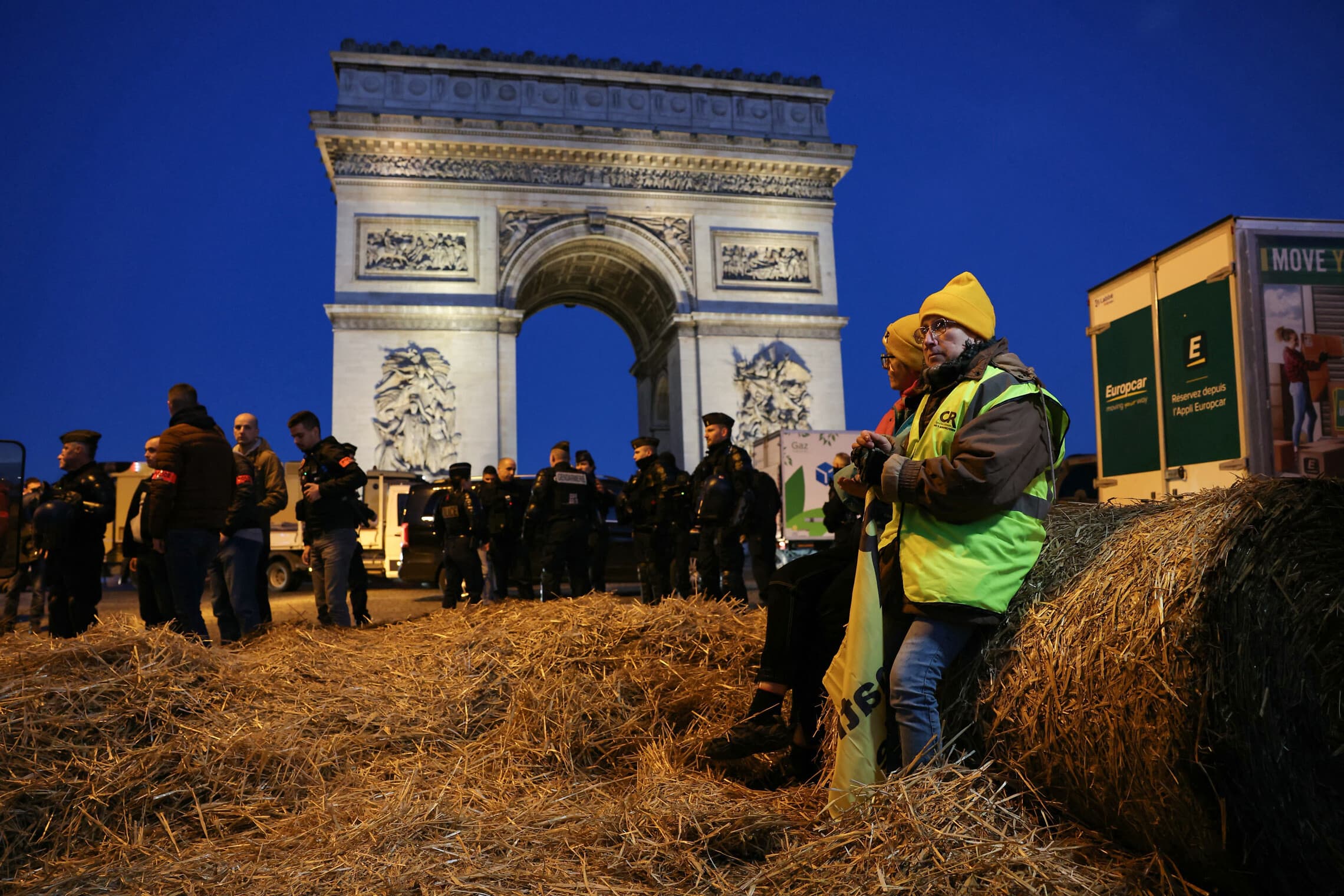 חקלאים צרפתים יושבים על חבילת חציר בזמן ששוטרים מאבטחים את האזור מול שער הניצחון ליד שדרת השאנז אליזה במהלך הפגנה של איגוד האיכרים הצרפתי בפריז ב-1 במרץ 2024 (צילום: Thomas SAMSON / AFP)