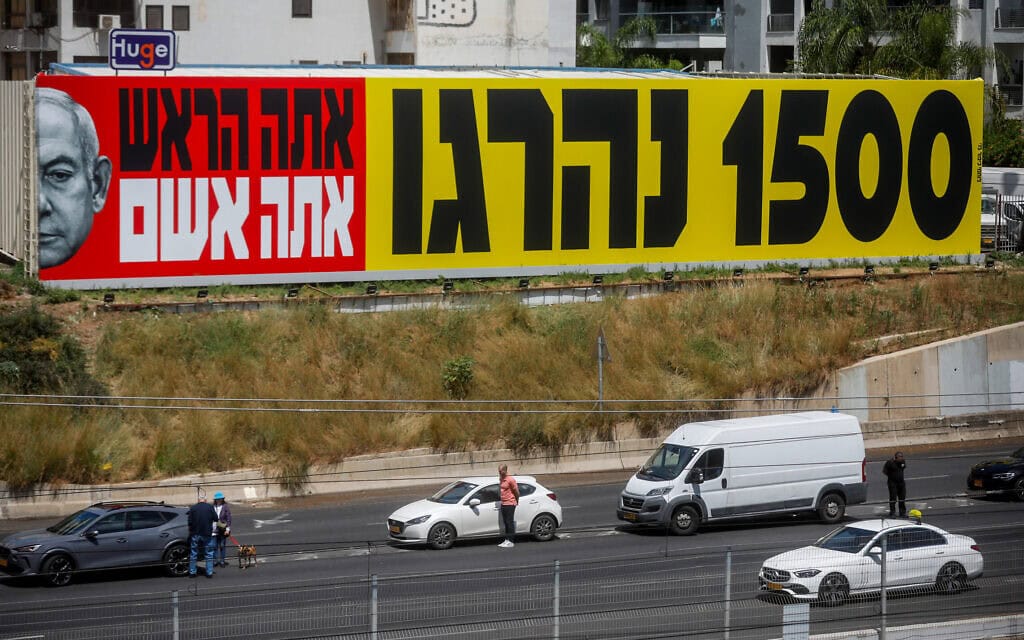 ישראלים עומדים בצפירה באיילון לצד שלט נגד ראש הממשלה בנימין נתניהו. 13 במאי 2024 (צילום: מרים אלסטר/פלאש90)