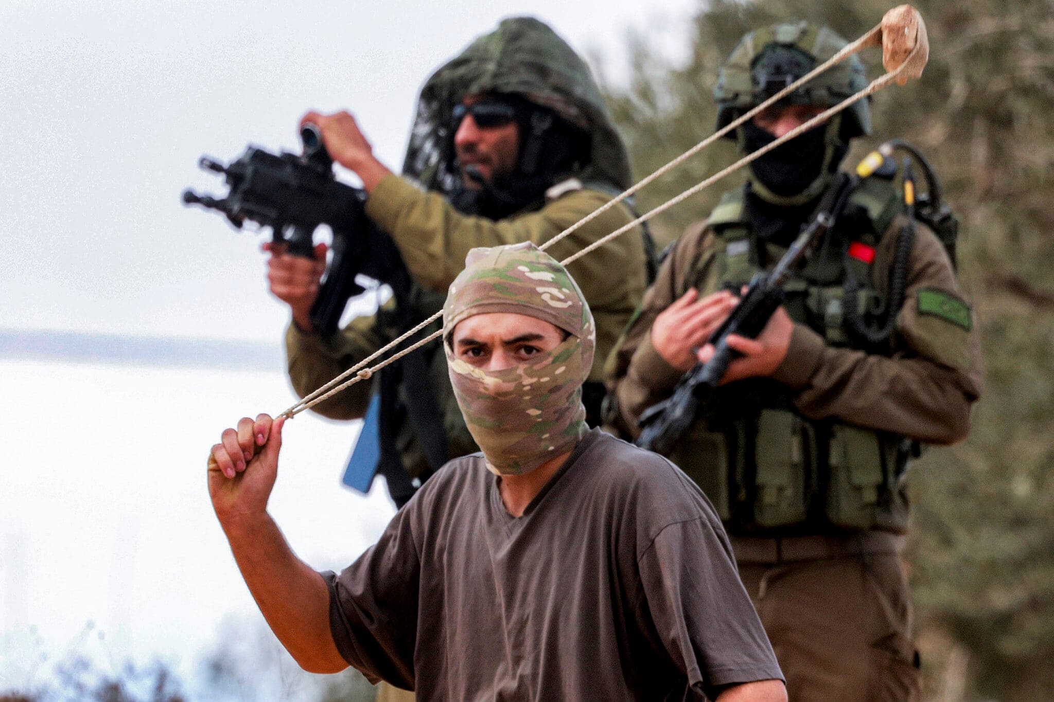 חיילי צה”ל עומדים מאחורי מתנחל רעול פנים המניף רוגטקה ומשליך אבנים לעבר פלסטינים שהתאספו למסיק הזיתים השנתי, במהלך תקיפת מתנחלים ישראלים על הכפר ביתא שבגדה המערבית, מדרום לשכם, ב-10 באוקטובר 2025 (צילום: Jaafar Ashtiyeh/AFP) חיילי צה”ל עומדים מאחורי מתנחל רעול פנים המניף רוגטקה ומשליך אבנים לעבר פלסטינים שהתאספו למסיק הזיתים השנתי, במהלך תקיפת מתנחלים ישראלים על הכפר ביתא שבגדה המערבית, מדרום לשכם, ב-10 באוקטובר 2025 (צילום: Jaafar Ashtiyeh/AFP)