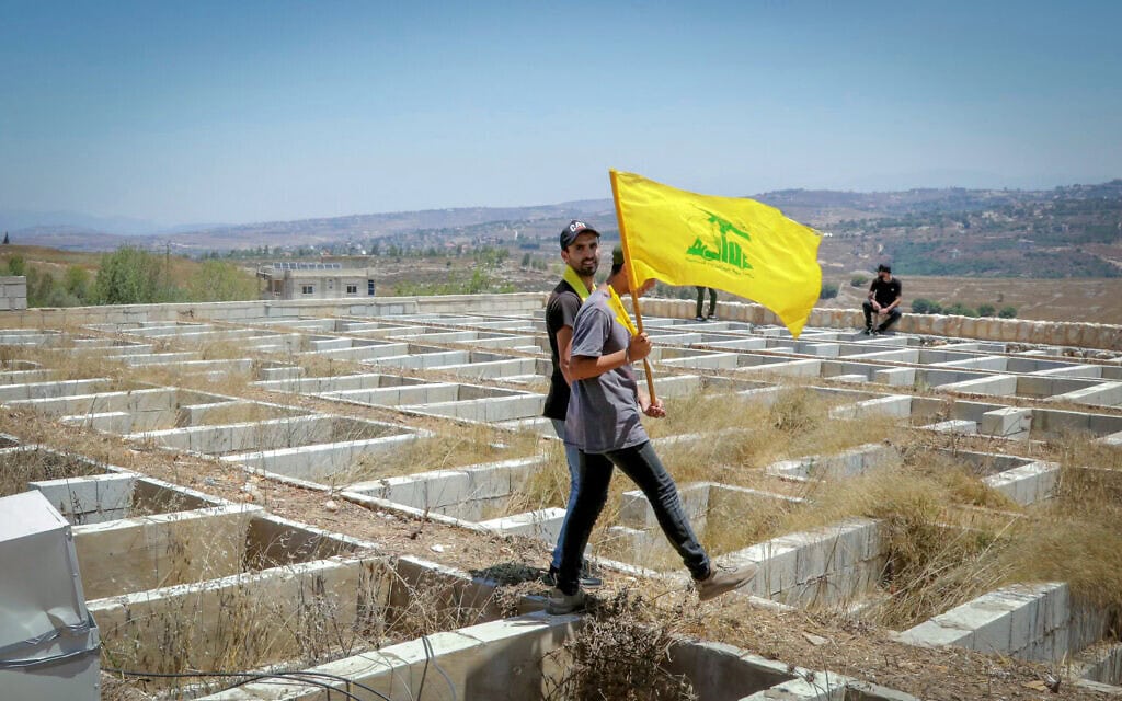 גבר מניף את דגל חזבאללה וצועד מעל קברים פתוחים במהלך הלוויה המונית בכפר מג'דל סלם שבדרום לבנון. 18 ביולי 2026 (צילום: Ibrahim AMRO / AFP) גבר מניף את דגל חזבאללה וצועד מעל קברים פתוחים במהלך הלוויה המונית בכפר מג'דל סלם שבדרום לבנון. 18 ביולי 2026 (צילום: Ibrahim AMRO / AFP)