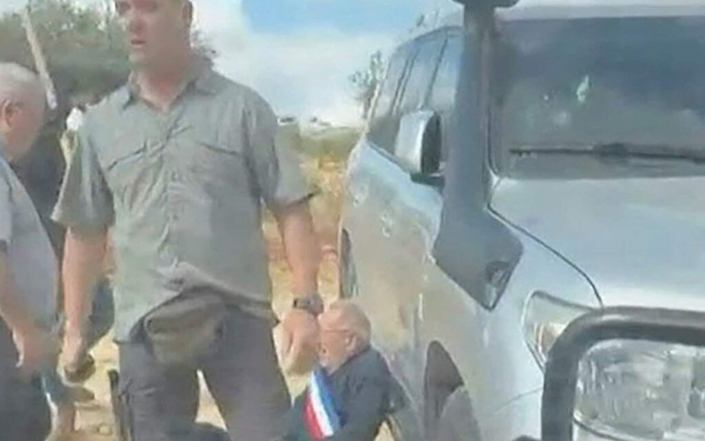צילום מסך מתוך תיעוד שהופץ ברשתות החברתיות מציג גבר עוטה אבנט כחול-לבן-אדום, מהסוג המשמש נבחרי ציבור בצרפת, נשען על כלי רכב בעוד רעולי פנים חוסמים ומכתרים מכונית שבה נסעו פעילים בכפר הנוצרי טייבה שבגדה המערבית. 17 באוקטובר 2026 (צילום: UGC / AFP)